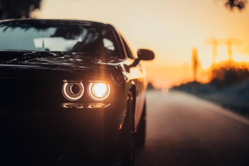 A sleek black car with bright headlights on a road at sunset